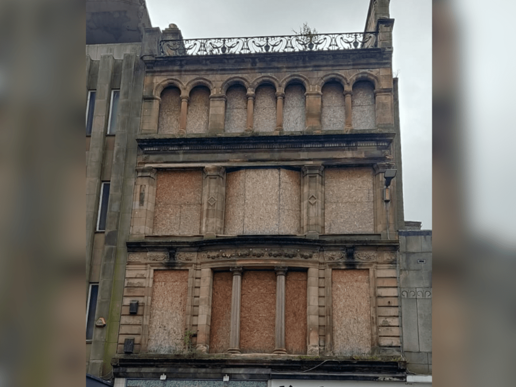 A photo of an above-shop flat in a high street in Glasgow. The building, apart from the shop, is entirely boarded up.