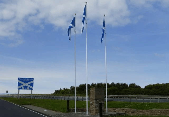 A photo of the Anglo-Scottish border, from the English side, looking towards the Welcome to Scotland sign. Three saltire flags on flagpoles in the centre.