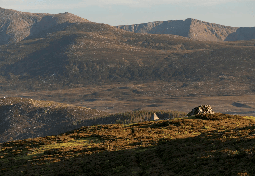 A stock photo of the Scottish Highlands, largely devoid of people or wildlife apart from some sitka spruce plantations, a cairn and what may be the top of the roof of a house.